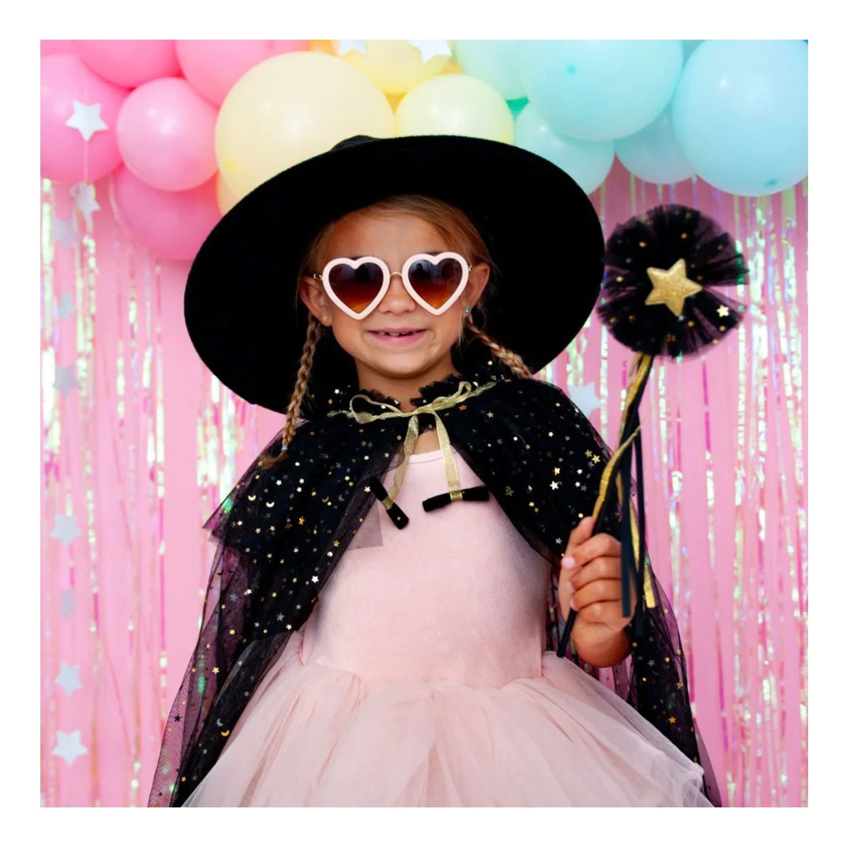 A child wearing Sweet Wink’s black Tulle witch cape with glitter stars that ties at the front neck, holding a black glitter star tulle wand w/ a gold star in center to match cape & Black Witch Hat Set.